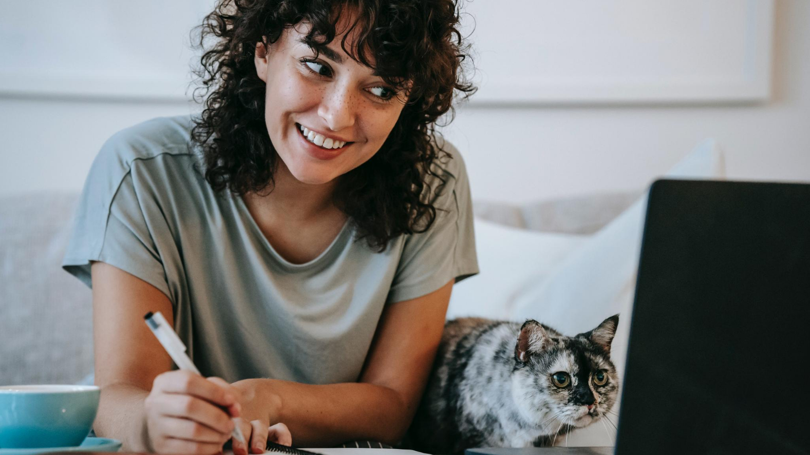 glad woman with cat brainstorming and writing in a planner while using laptop