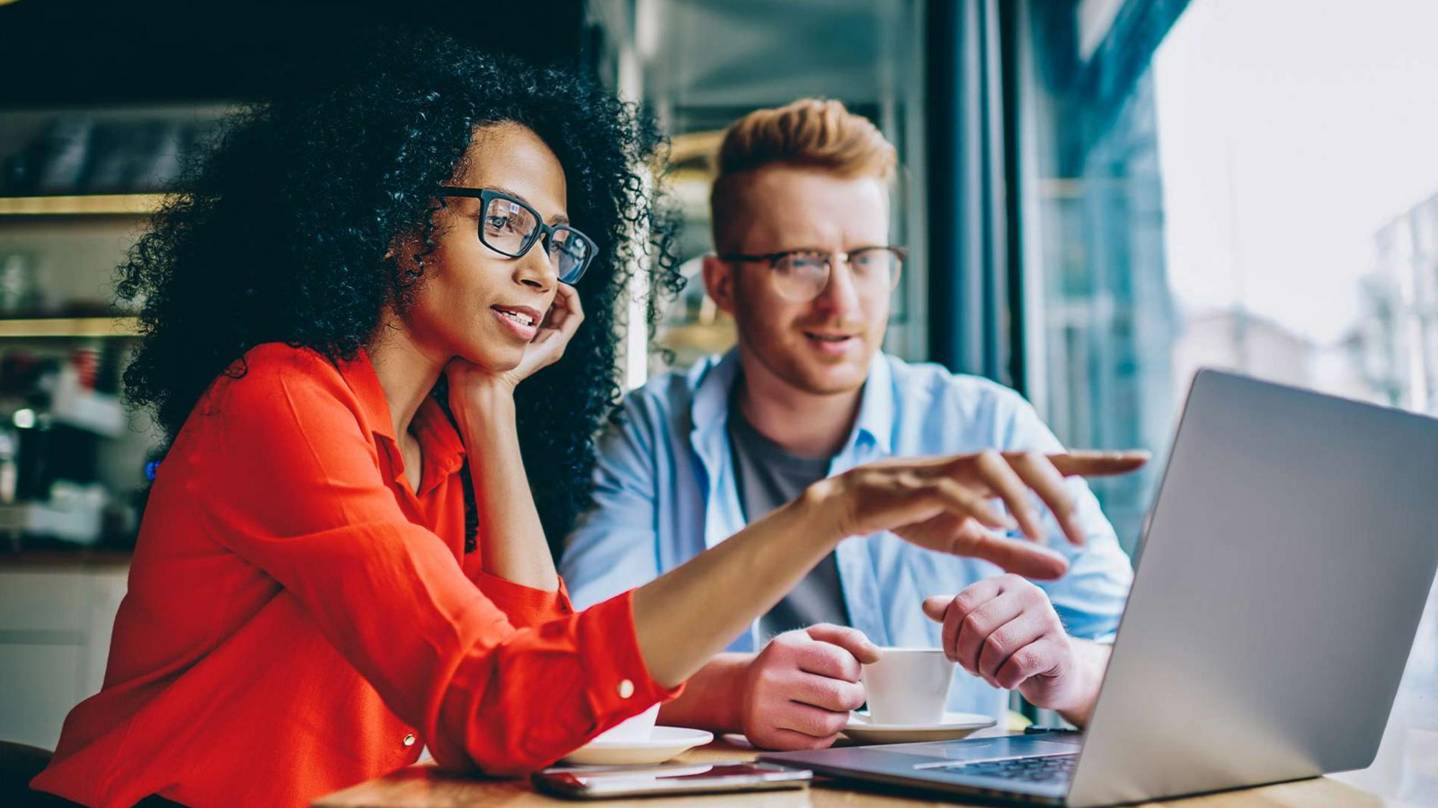 female points to screen on laptop while discussing onsite optimization project with colleague in coffee shop