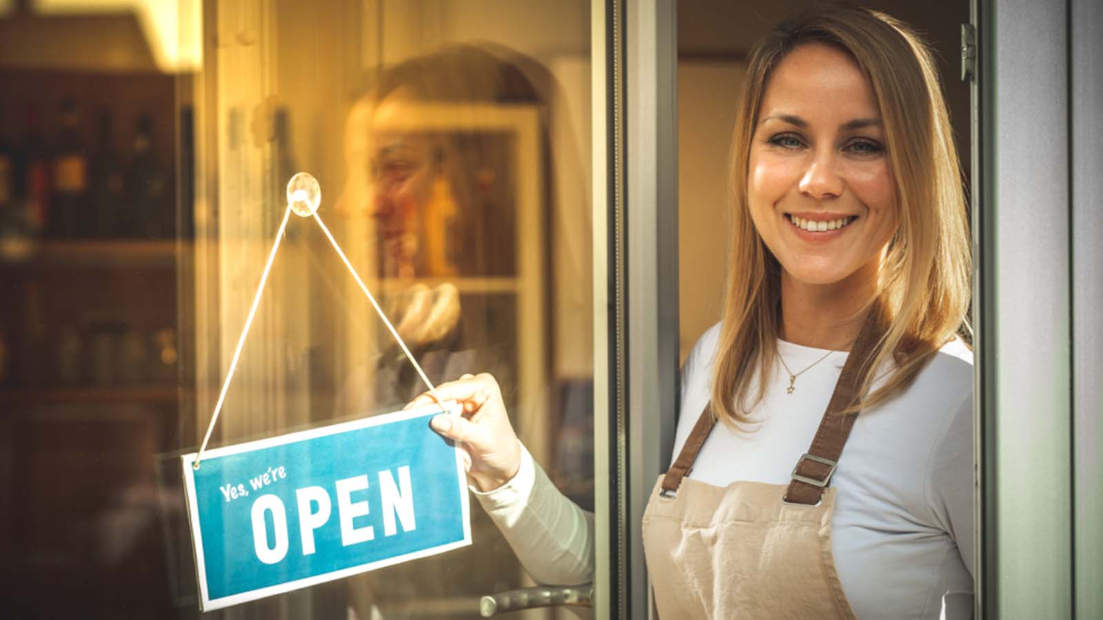 coffee shop business owner wearing an apron and holding the sign ''we are open'' on the door